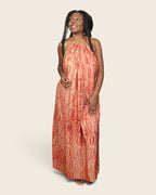 A woman modeling an Adire sleeveless, halter-neck maxi dress with an orange and beige paisley pattern, set against a solid light beige background.
